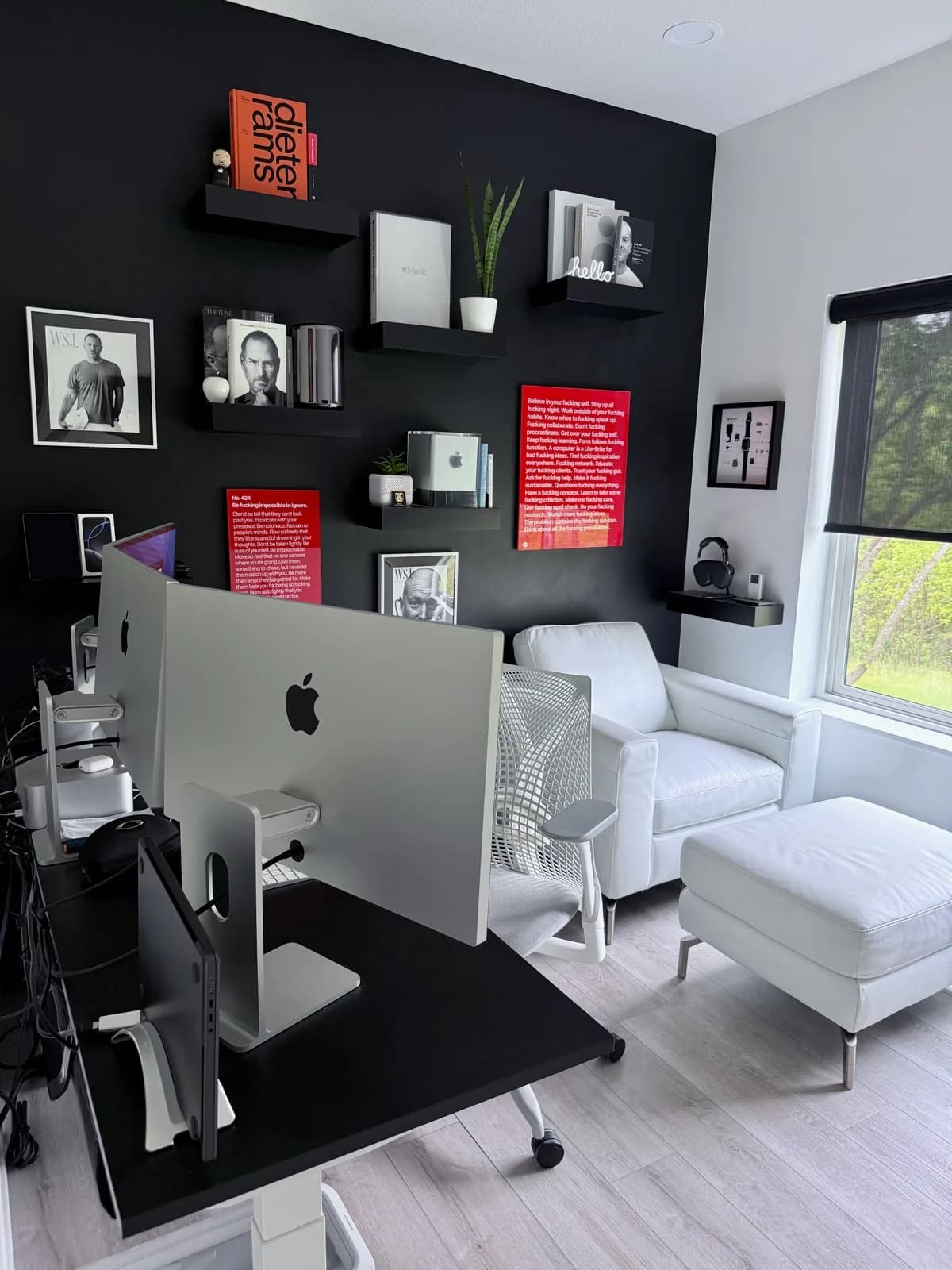 Justin Bartak's workspace: an iMac on a dark desk facing a matte black accent wall arranged with a Dieter Rams book, framed photographs of Steve Jobs, a Power Mac G4 Cube, and two red essay posters. A white leather lounge chair and ottoman sit beside a window with daylight and outdoor greenery.